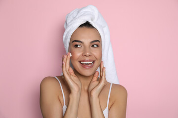 Beautiful young woman with towel applying cream on face against pink background