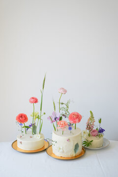 Close-up of creative vegan desserts. Three white beautiful wedding cake with cream and colored fresh flowers on a white table with a tablecloth and a white background