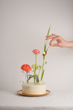 A small white cake decorated with flowers with a hand reaching for it a golden spoon on a white background