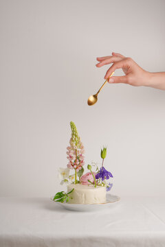 White cake for one person decorated with flowers with lupine with a hand reaching for it a golden spoon on a white background