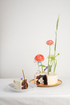 White cake with ranunсulus with a chocolate cut on a gold plate on a white table with a piece with a spoon next to
