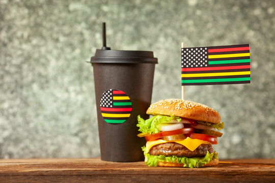 Classic American burger with alternative African American juneteenth flag on the top and black paper cup with straw over wooden background. Close-up with selective focus.