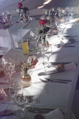 Dreamy table setting of a wedding dinner with an arrangement of glasses, flowers, napkins, multiple items of cutlery, and small burning candles. Sun shining on the table. Love and happiness. 