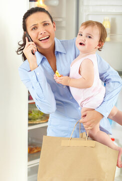 Too Much To Juggle Sometimes. An Overwhelmed Working Mom Speaking On The Phone While Holding Her Baby And A Bag Of Groceries.