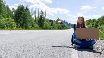 A young girl is sitting on the road with a sign in her hands, hitchhiking around the country. The girl is trying to catch a passing car for a trip. A girl with a backpack went on vacation hitchhiking