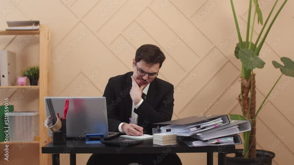Poster office worker man with thoughtful look, writes something down in notebook while sitting at table wit