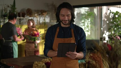 Young male staff wearing apron holding digital tablet standing in flower shop service