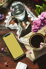 Aesthetic lifestyle flat lay, wireless headphones, mockup screen of phone, lilac flowers decorations, cup of coffee and glass of water.Listening music or audiobook, surfing phone lifestyle