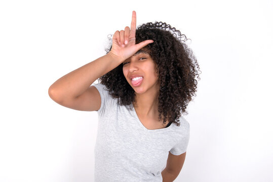 Funny Young beautiful girl with afro hairstyle wearing grey t-shirt over white wall makes loser gesture mocking at someone sticks out tongue making grimace face.
