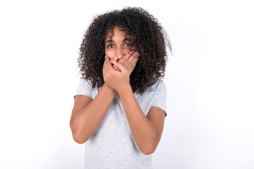 Fototapeta na wymiar Stunned Young beautiful girl with afro hairstyle wearing gray t-shirt over white background covers both hands on mouth, afraids of something astonishing