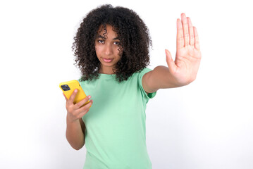 Young beautiful girl with afro hairstyle wearing green t-shirt over white wall using and texting with smartphone with open hand doing stop sign with serious and confident expression, defense gesture