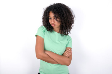 Gloomy dissatisfied Young beautiful girl with afro hairstyle wearing green t-shirt over white background looks with miserable expression at camera from under forehead, makes unhappy grimace