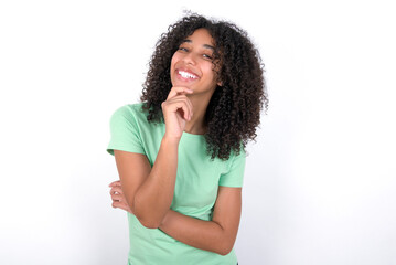 Fototapeta na wymiar Young beautiful girl with afro hairstyle wearing green t-shirt over white background laughs happily keeps hand on chin expresses positive emotions smiles broadly has carefree expression