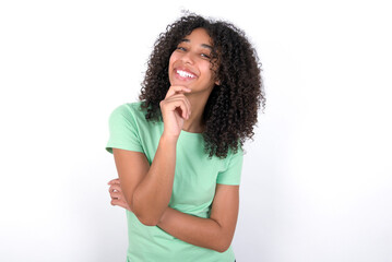 Young beautiful girl with afro hairstyle wearing green t-shirt over white background laughs happily keeps hand on chin expresses positive emotions smiles broadly has carefree expression