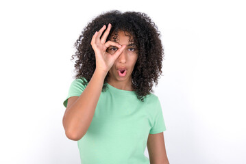 Young beautiful girl with afro hairstyle wearing green t-shirt over white background doing ok gesture shocked with surprised face, eye looking through fingers. Unbelieving expression.