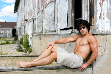 Sexy athletic young male sits casually outside in front of a broken down barn on a farm in Iowa.  