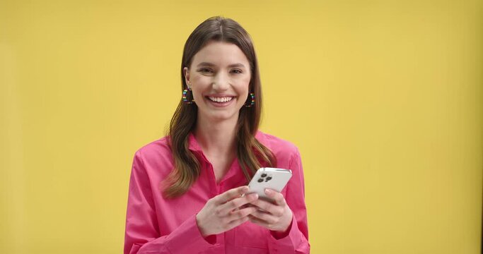 Close up of joyful beautiful Caucasian female in pink blouse typing on mobile phone searching internet using social network and smiling looking at camera standing on yellow background wall, portrait