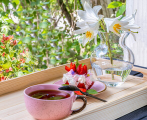 Teacup of tea with dessert. Pink teacup, pink saucer, white lily, strawberry. Summer garden, wooden tray. Food and drink