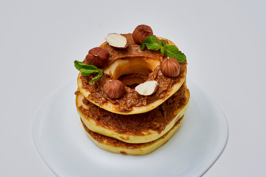 Apple rounds dialed on top of each other with peanut butter, almonds and nuts, on white serving plate. Healthy food