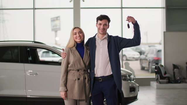 portrait of happy male and female buyers with keys to new automobile in their hands, rejoice at successful deal stand backdrop of vehicle in auto dealership, smile and look at camera
