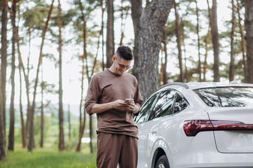 Young attractive smiling man with beard standing near his car and holding mobile phone