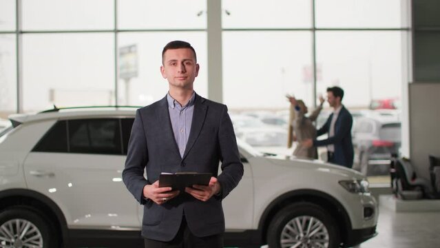 car sales center, portrait of a male sales manager background of happy owners of a new automobile in an auto showroom, smiling and looking at camera