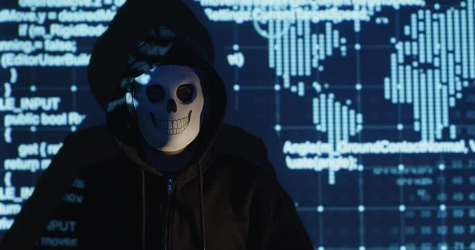 A man in a dark jacket and a protective mask on his face sits against the background of computer code graphics and a world map. Hacker hides his face for anonymity.