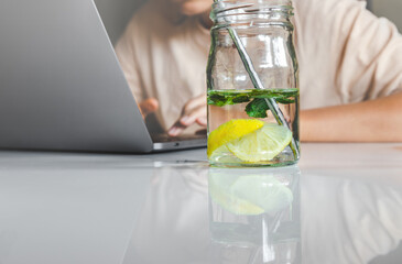 Water with lemon and mint leaves in glass jar and female person typing on laptop in background.