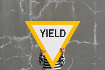 Person hidden holding large yield road sign. Bright yellow against cracked gray wall. - obrazy, fototapety, plakaty