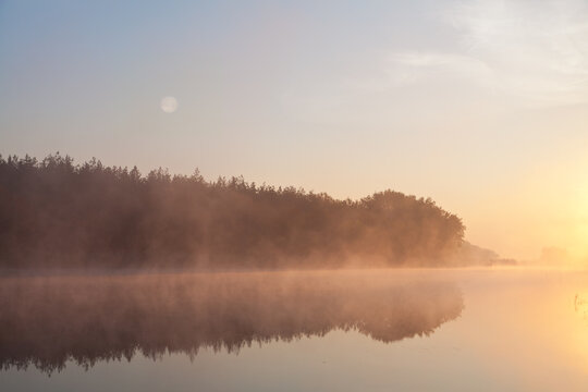 Foggy morning over calm river, pink fog against morning sun, full moon. Ukraine, peace.