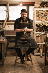 Vertical playful male carpenter pressing in hands wood shaving chips, sitting on chair after...