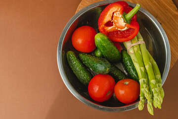 Vegetables lie in a metal bowl: tomatoes, asparagus, cucumbers, red bell peppers. on a wooden board and brown background. place for text. view from above