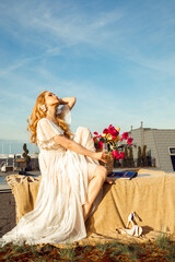 Vertical of attractive curly blond woman with close eyes in wedding dress with basket of flowers and drink on roof top