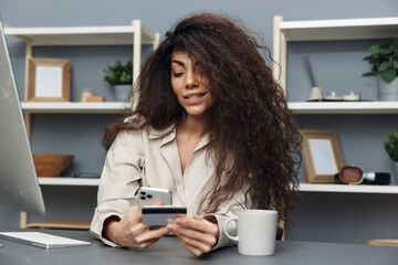 Cute cheerful tanned adorable curly Latin woman in linen shirt admiring her credit card in home office interior. Copy space Mockup Banner. Shopaholic lady uses modern computer for online shopping
