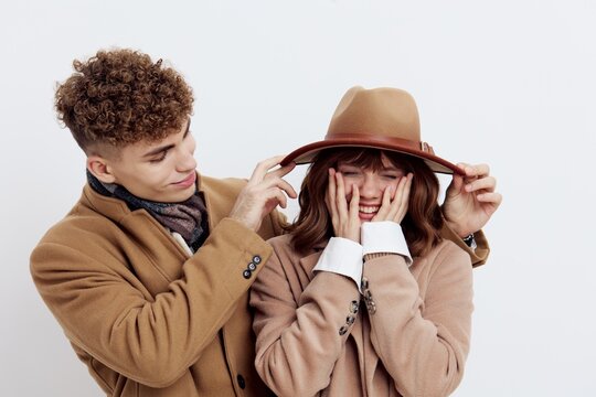 a cute couple stands on a light background in an autumn coat and sweaters. a man is standing behind her, looking tenderly at the woman, helping her to straighten her hat while she put her fingers on