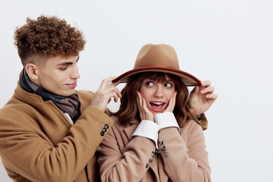 a cute couple stands on a light background in an autumn coat and sweaters. a man is standing behind her, looking tenderly at the woman, helping her to straighten her hat while she put her fingers on