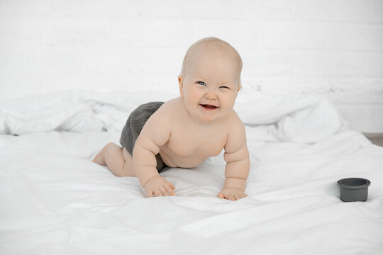 Cheerful little baby crawl on big bed, have fun and laugh on white background. Portrait of beautiful naked infant child, free copy space. Concept of happy childhood and caring for children