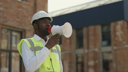 Serious african american engineer using megaphone to communicate with workers at construction site. Concept of people, professionalism and building industry. - Powered by Adobe