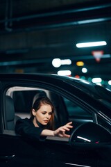 vertical photo from the side, at night, of a woman sitting in a black car and looking out of the window and reaching out to the side view mirror to correct it