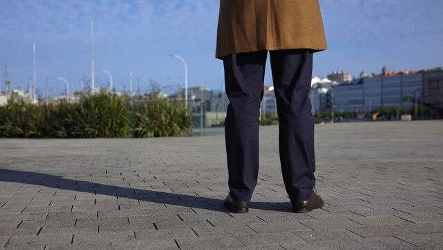 Men's feet in dark blue trousers and brown classic and shoes. Rear view. Part of the brown business polto is visible. The legs stand on the asphalt and then begin to move forward. The area is paved