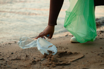 Cropped photo of African woman picking up spilled trash garbage from sand on beach in green plastic bag. Womans hand cleaning up used plastic bottles. Ecology, environmental conservation, pollution. 