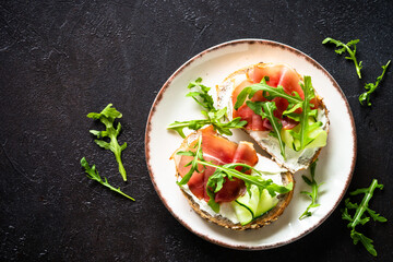 Open sandwich with cream cheese and jamon with arugula in white plate. Top view at dark table.