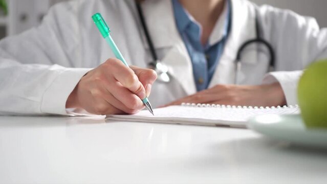 A woman doctor sits at her desk writes with a pen in ink filling out the documentation of a medical history card for patients, issuing a prescription for buying medicines for a patient
