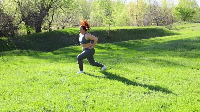 Young overweight woman in fitness suit jump squat with one leg forward standing on green lawn in park on sunny day, slow motion. Healthy lifestyle, exercise and fitness outdoors. Sports activity.