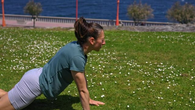 The girl does the exercise, lying on the grass, overlooking the ocean. A girl in a green T-shirt and gray shorts. The girl kneels and leans on outstretched straight arms, then alternately touches the