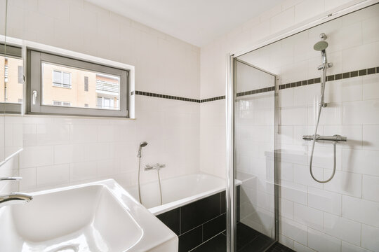 Modern Bathroom Interior Design With White Ceramic Tiles With A Bathtub Under The Window, Glass Shower, White Sink And Black Tiled Floor.