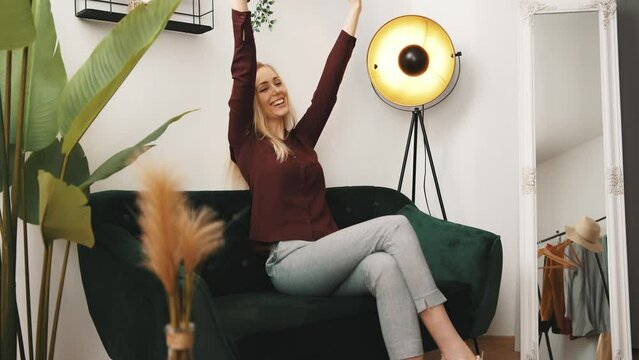 Success. Young long-haired blonde caucasian girl throws her arms in the air while sitting on a dark green couch to show her positive reaction after finding out about her promotion. High quality 4k