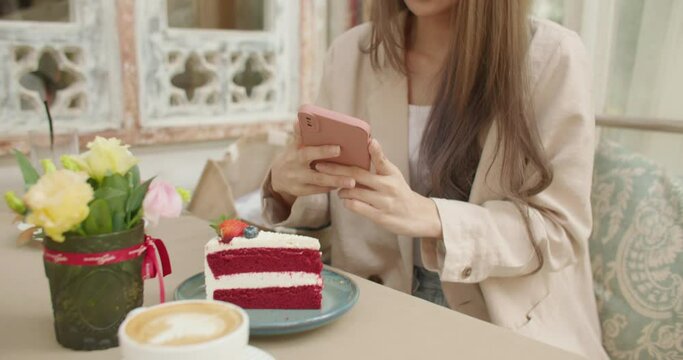Close up of woman holds smartphone taking pictures of sweet dessert and coffee to share photos on social media. Attractive satisfied young girl feeling pleasure with delicious food cake at coffeeshop.
