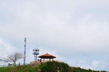 A red-roofed gazebo and electrical tower building on top of a hill under a large blue sky and white...