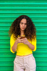 Portrait of pretty brazilian girl using smartphone - Elegant latin woman from Brazil with curly hair and dressed in yellow and isolated on green - Brazilian flag colours.